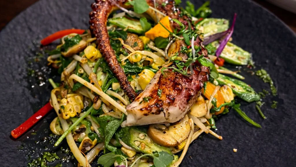 Close view of grilled octopus over vegetables on a black plate