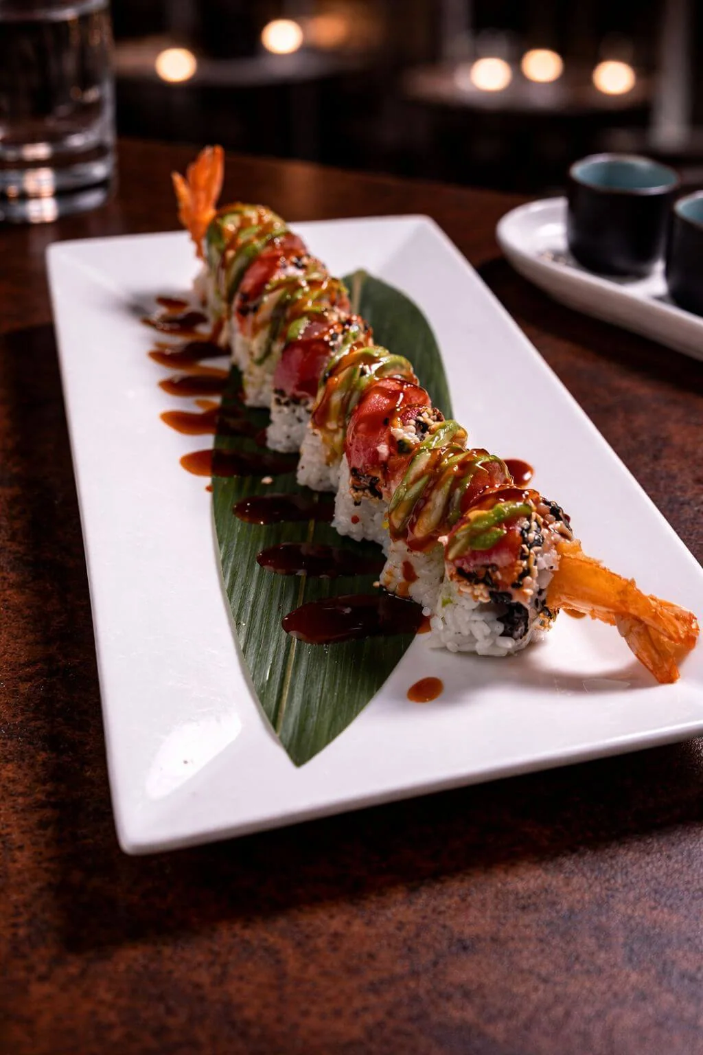 Sushi roll topped with tuna and dark glaze, with a tempura shrimp tail, on a white plate